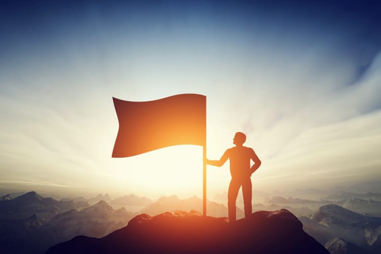 Proud man raising a flag on the peak of the mountain. Successful challenge concept, new achievement